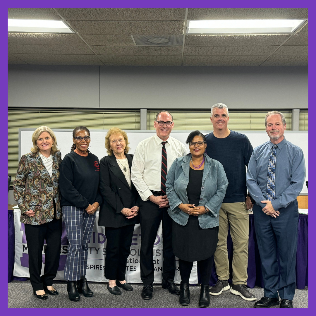 Photo shows seven adults standing next to each other in meeting room